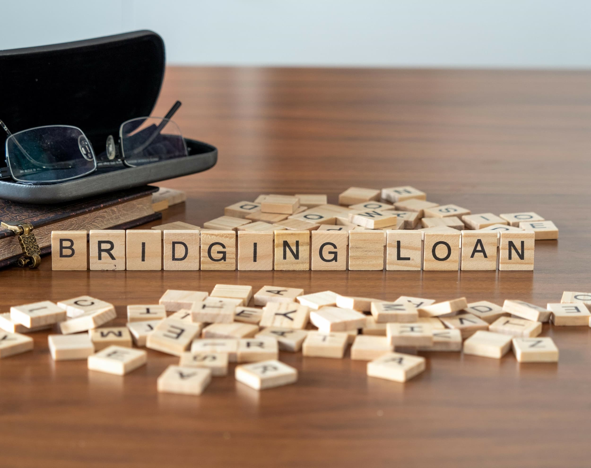 Wooden letter tiles spell out ‘bridging loan’ on a desk with reading glasses and a notebook nearby.