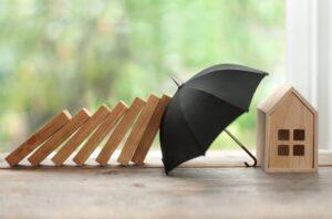 A black umbrella prevents several wooden blocks from falling onto a small wooden home, representing mortgage protection.