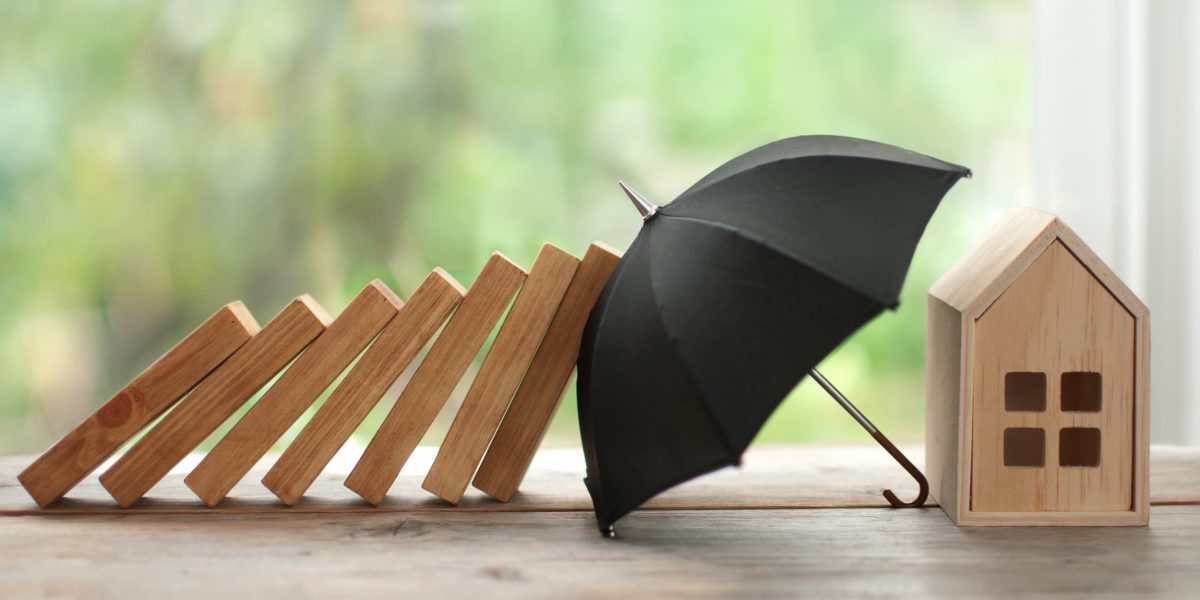 A black umbrella prevents several wooden blocks from falling onto a small wooden home, representing mortgage protection.